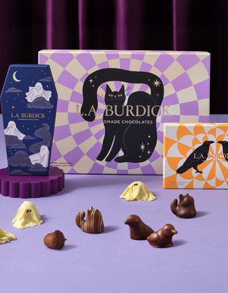 Assorted artisan chocolates shaped like ghosts, acorns, cats, and birds displayed in front of three colorful L.A. Burdick chocolate boxes with playful designs.