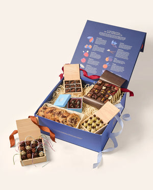An open blue box with an assortment of gourmet chocolates (chocolate mice, Snowmen, deer and assorted shapes) and baked goods (chocolate chip cookies). The main box is navy blue the inside of the open lid depicts rows of colorful illustrations and text.