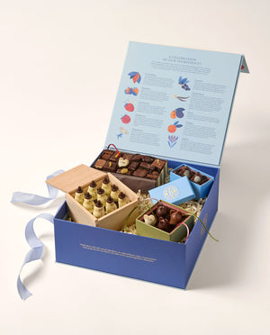 An open dark and light blue box containing an assortment of gourmet chocolates (chocolate mice, Snowmen, penguins and assorted shapes). The outside of the box is navy blue the inside lid of the open is light blue with rows of colorful illustrations and text.