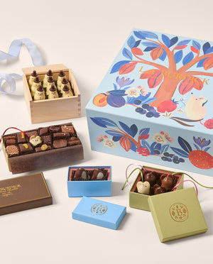 A beautifully arranged assortment of gourmet chocolates in elegant packaging, including a large illustrated blue gift box, a wooden box of white chocolate snowmen, a dark chocolate box, and smaller blue and green boxes with chocolate penguins and mice, all on a light beige background.
