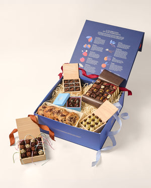 An open blue box with an assortment of gourmet chocolates (chocolate mice, Snowmen, deer and assorted shapes) and baked goods (chocolate chip cookies). The main box is navy blue the inside of the open lid depicts rows of colorful illustrations and text.