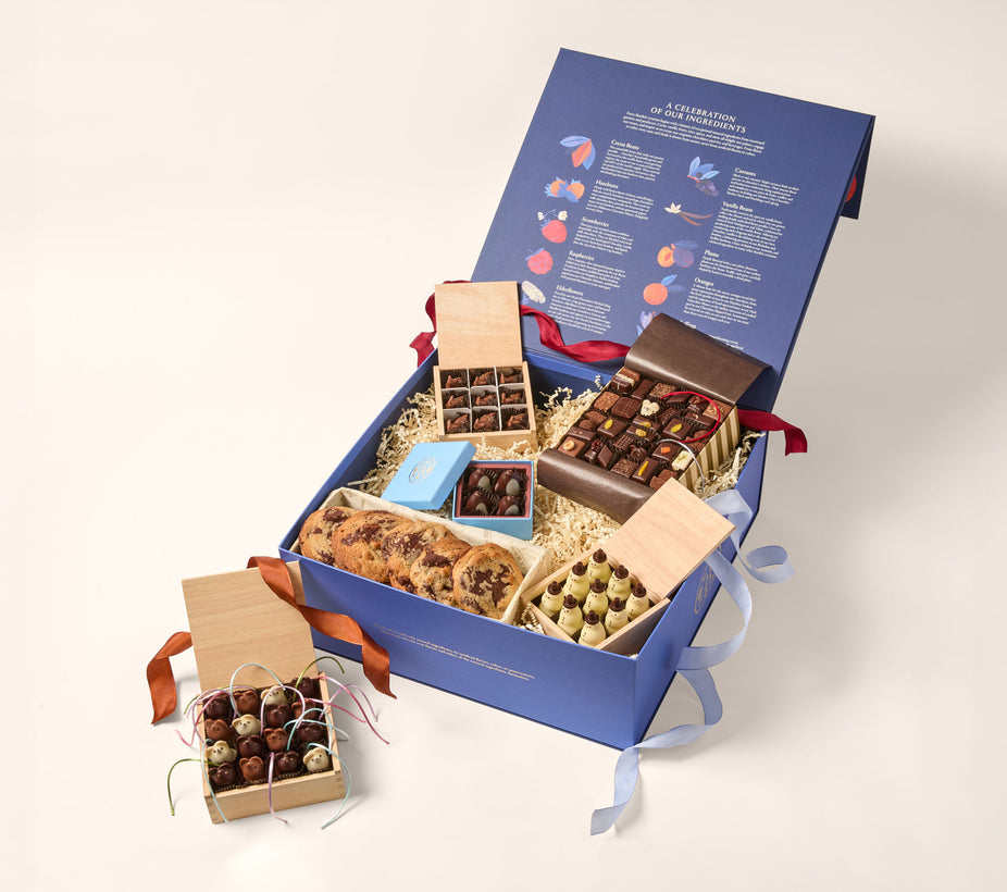 An open blue box with an assortment of gourmet chocolates (chocolate mice, Snowmen, deer and assorted shapes) and baked goods (chocolate chip cookies). The main box is navy blue the inside of the open lid depicts rows of colorful illustrations and text.