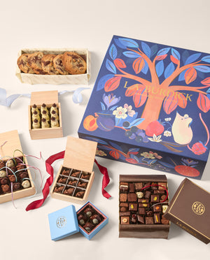 An assortment of gourmet chocolates ( chocolate mice, Snowmen, deer and assorted shapes) and baked goods (chocolate chip cookies) arranged around a large decorative gift box. The main box is navy blue with a colorful tree illustration and a mouse motif.