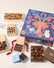 An assortment of gourmet chocolates ( chocolate mice, Snowmen, deer and assorted shapes) and baked goods (chocolate chip cookies) arranged around a large decorative gift box. The main box is navy blue with a colorful tree illustration and a mouse motif.