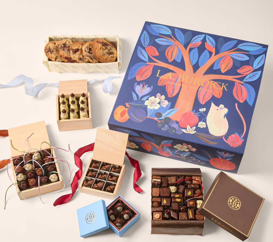 An assortment of gourmet chocolates ( chocolate mice, Snowmen, deer and assorted shapes) and baked goods (chocolate chip cookies) arranged around a large decorative gift box. The main box is navy blue with a colorful tree illustration and a mouse motif.