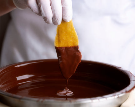 A dried mango is shown half-dipped in melted chocolate, being held by one of our chocolatiers.
