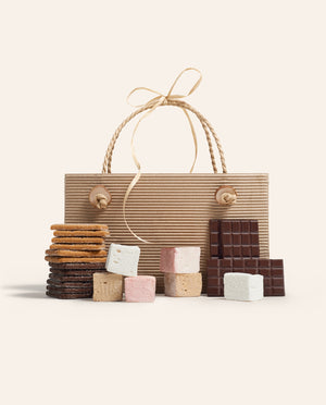 Bag with the components to make s’mores in front, gram crackers, marshmallows and chocolate bars on a cream background.