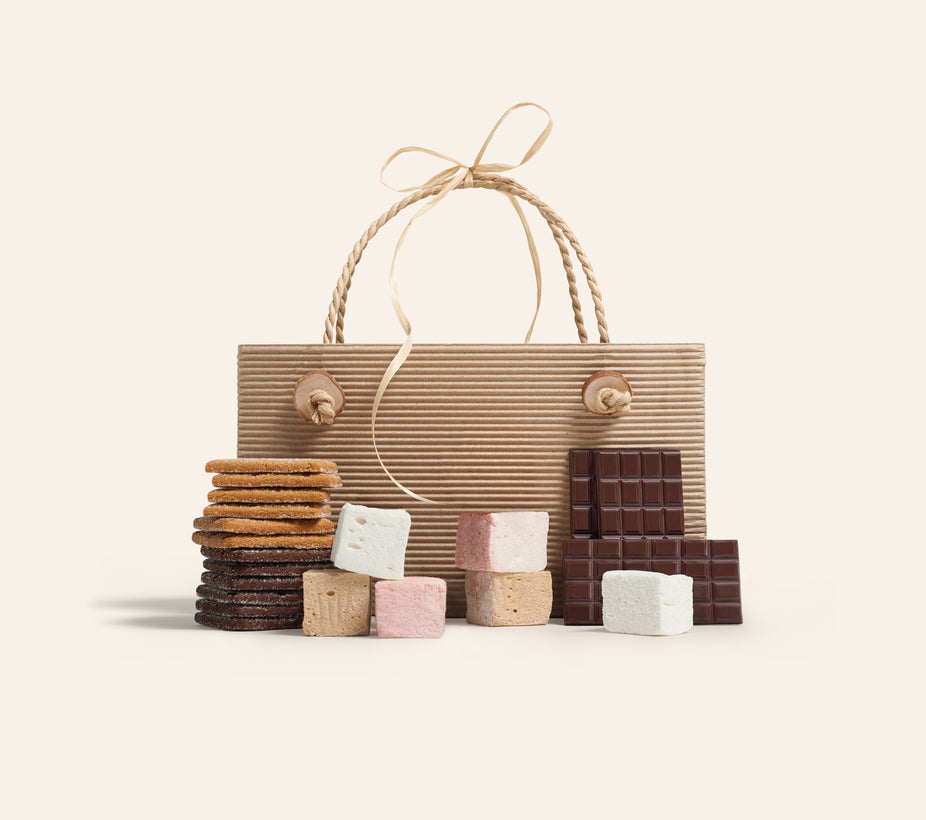 Bag with the components to make s’mores in front, gram crackers, marshmallows and chocolate bars on a cream background.