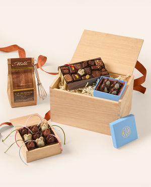 A gourmet chocolate gift set arranged in a light wood box with shredded paper filler, alongside a small blue box lid, a bag of milk chocolate pieces, and a whisk, all on a clean off-white background.