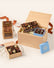 A gourmet chocolate gift set arranged in a light wood box with shredded paper filler, alongside a small blue box lid, a bag of milk chocolate pieces, and a whisk, all on a clean off-white background.