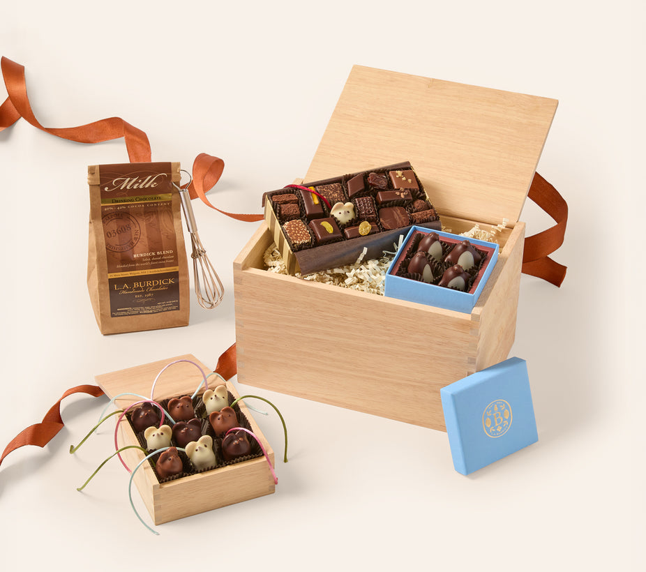 A gourmet chocolate gift set arranged in a light wood box with shredded paper filler, alongside a small blue box lid, a bag of milk chocolate pieces, and a whisk, all on a clean off-white background.