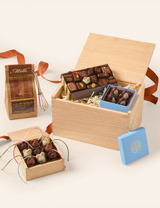 A gourmet chocolate gift set arranged in a light wood box with shredded paper filler, alongside a small blue box lid, a bag of milk chocolate pieces, and a whisk, all on a clean off-white background.