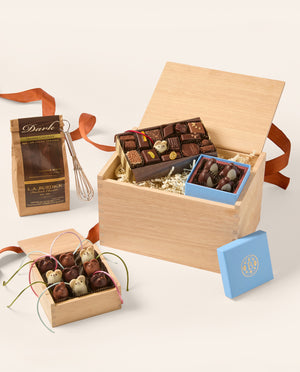 A gourmet chocolate gift set arranged on a light beige background. The set includes a large open wooden box filled with assorted chocolates, a smaller blue box of chocolates, a brown paper bag labeled 'Dark' with a whisk, and a small open wooden box of decorated chocolates tied with ribbons.