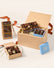 A gourmet chocolate gift set arranged on a light beige background. The set includes a large open wooden box filled with assorted chocolates, a smaller blue box of chocolates, a brown paper bag labeled 'Dark' with a whisk, and a small open wooden box of decorated chocolates tied with ribbons.