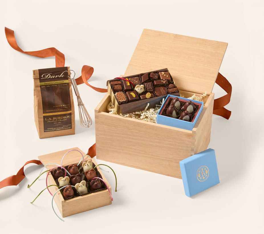 A gourmet chocolate gift set arranged on a light beige background. The set includes a large open wooden box filled with assorted chocolates, a smaller blue box of chocolates, a brown paper bag labeled 'Dark' with a whisk, and a small open wooden box of decorated chocolates tied with ribbons.