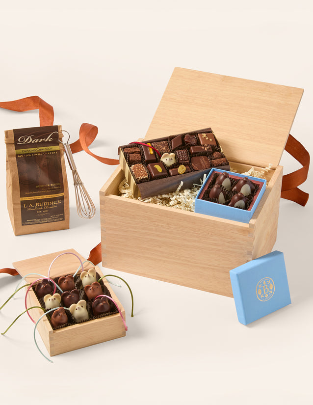 A gourmet chocolate gift set arranged on a light beige background. The set includes a large open wooden box filled with assorted chocolates, a smaller blue box of chocolates, a brown paper bag labeled 'Dark' with a whisk, and a small open wooden box of decorated chocolates tied with ribbons.