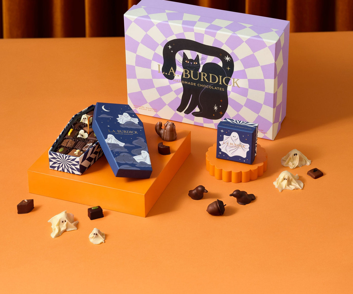 A Halloween-themed arrangement of L.A. Burdick chocolates displayed on an orange surface with decorative packaging featuring a black cat silhouette and assorted handcrafted chocolate pieces scattered around.