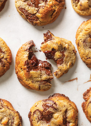 Close-up of freshly baked artisan chocolate chip cookies arranged on a white surface, with one cookie broken in half revealing melted chocolate inside.
