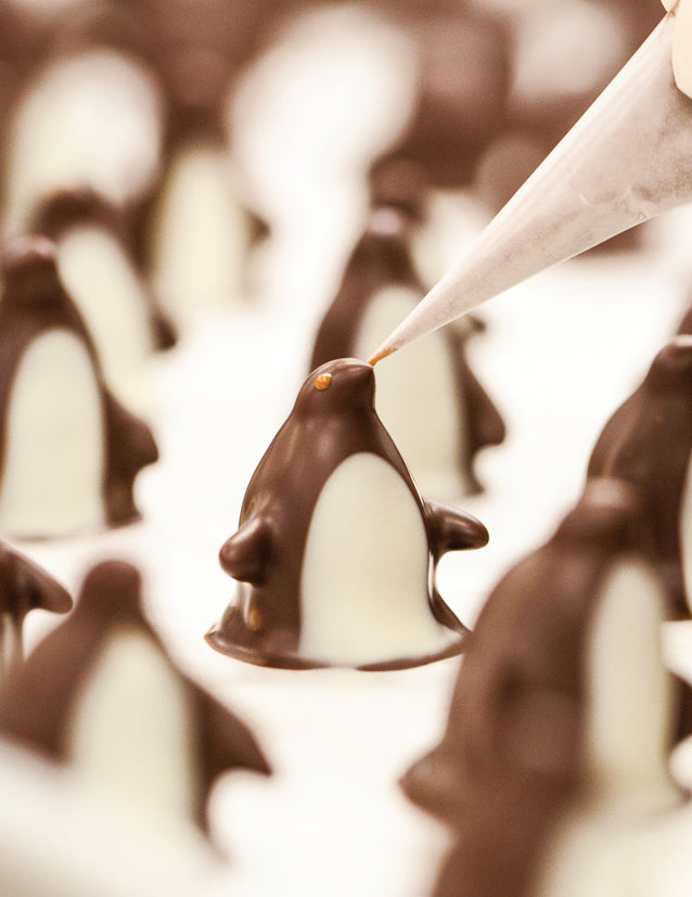 Rows of dark chocolate penguins with white chocolate bellies on a white surface, The center penguin is having a milk chocolate eye added with a piping tip.