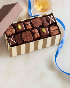 An open striped gift box containing an assortment of artisan handmade chocolates with various toppings, next to a golden ribbon and a glass of amber liquid.