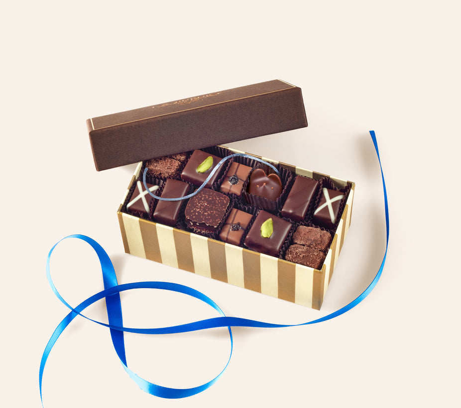 A box of assorted chocolate bonbons including a dark chocolate mouse  with a blue silk tail. Below the box is a blue ribbon. 