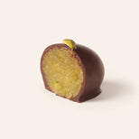 Chocolate covered pistachio marzipan egg cut in half to show the slightly green interior, topped with an pistachio bit, on a cream background