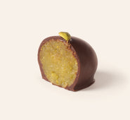Chocolate covered pistachio marzipan egg cut in half to show the slightly green interior, topped with an pistachio bit, on a cream background