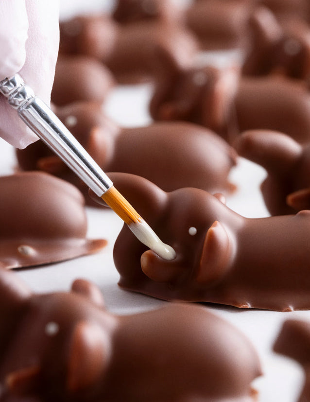 Chocolate elephants being decorated with a paintbrush on a white surface.