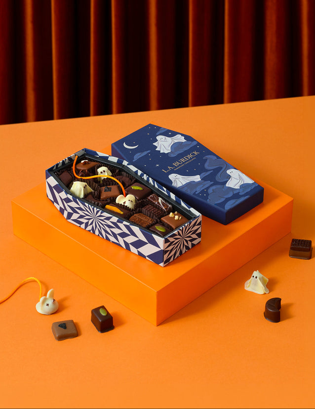 A boxed assortment of chocolates displayed on an orange pedestal against a dark orange curtain background. The gift box is open, showing assorted dark and milk chocolate pieces, with the closed lid placed beside it.