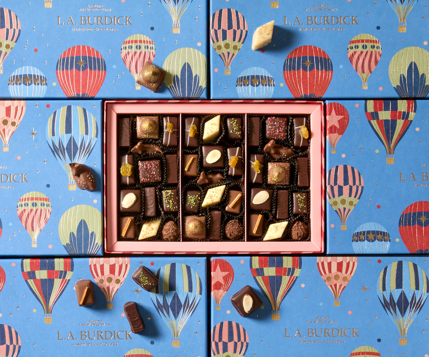 Box with assorted chocolates on a cream background. A light blue lid is adorned with vector illustrations of colorful hot air balloons and stars the center of the lid is embossed with a gold foil L.A. Burdick logo. The open box is striped dark pink and light pink on the sides and contains around 37 chocolates of various shapes and sizes adorned with various nuts and fruits.