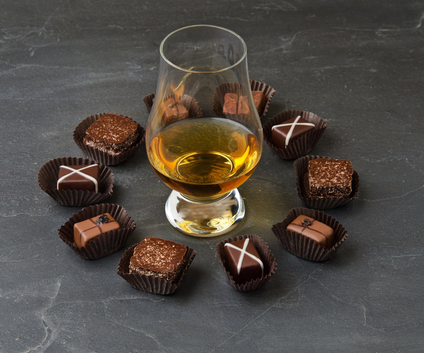 A glass of Scotch on a slate surface surrounded by various bonbons.