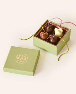 Four chocolate mice, dark chocolate, milk chocolate and white chocolate, with silken green and pink tails, in a square green box. The lid of the box is embossed with a gold foil L.A. Burdick logomark.