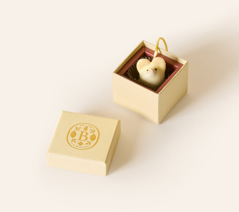White chocolate mouse with a yellow cream silken tail in a square cream box. The lid of the box is embossed with a gold foil L.A. Burdick logomark.