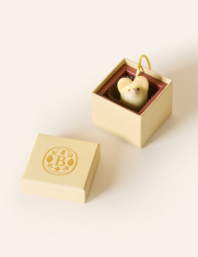 White chocolate mouse with a yellow cream silken tail in a square cream box. The lid of the box is embossed with a gold foil L.A. Burdick logomark.
