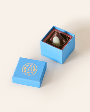 Dark chocolate penguin, with a white chocolate belly in a square light blue box. The lid of the box is embossed with a gold foil L.A. Burdick logomark.