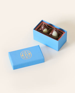 Two dark chocolate penguins with white chocolate bellys, in a rectangular light blue box. The lid of the box is embossed with a gold foil L.A. Burdick logomark.
