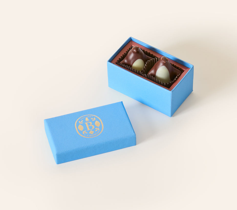 Two dark chocolate penguins with white chocolate bellys, in a rectangular light blue box. The lid of the box is embossed with a gold foil L.A. Burdick logomark.