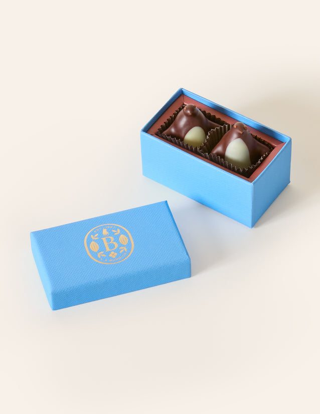 Two dark chocolate penguins with white chocolate bellys, in a rectangular light blue box. The lid of the box is embossed with a gold foil L.A. Burdick logomark.