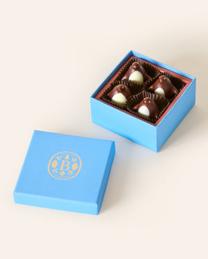 Four dark chocolate penguins with white chocolate bellys, in a square light blue box. The lid of the box is embossed with a gold foil L.A. Burdick logomark.