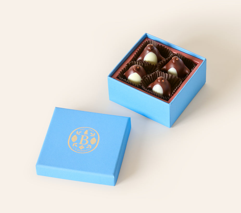 Four dark chocolate penguins with white chocolate bellys, in a square light blue box. The lid of the box is embossed with a gold foil L.A. Burdick logomark.