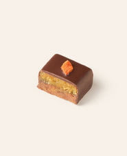 Square chocolate bonbon cut in half revealing a layered marzipan interior a orange carrot bit garnishes the top surface.